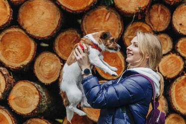 Smiling woman holding Jack Russell Terrier dog in front of wooden logs - VSNF00720