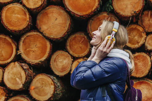 Lächelnde junge Frau hört Musik über Kopfhörer neben einem Stapel Holzscheite - VSNF00721