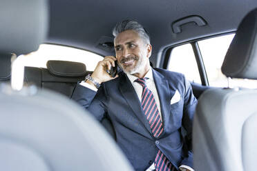 Lächelnder Geschäftsmann im Anzug, der im Auto mit seinem Smartphone telefoniert - JJF00790