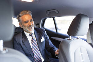 Smiling businessman wearing suit sitting on back seat in car - JJF00795