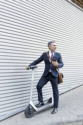 Businessman with smart phone and electric scooter standing near wall - JJF00816