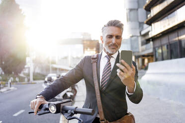 Geschäftsmann, der sein Smartphone im Stehen mit einem Elektroroller benutzt - JJF00824