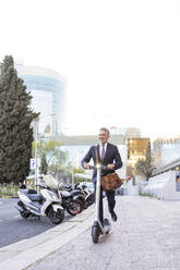 Smiling businessman riding electric scooter on sidewalk - JJF00826