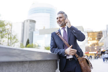 Smiling businessman talking on smart phone leaning on wall - JJF00832