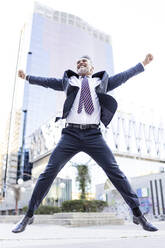 Happy businessman jumping with arms outstretched in front of building - JJF00838