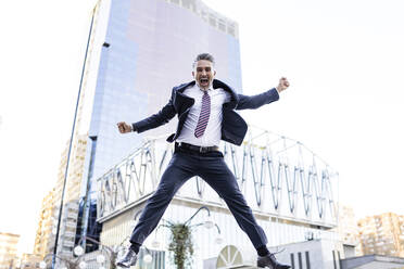 Excited businessman jumping in front of building - JJF00839