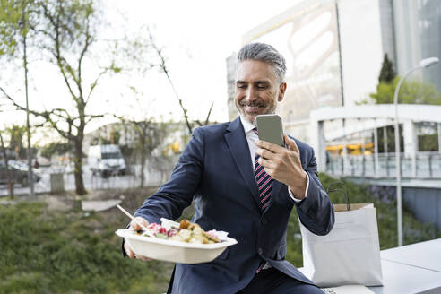 Businessman photographing meal box through smart phone sitting on wall - JJF00850