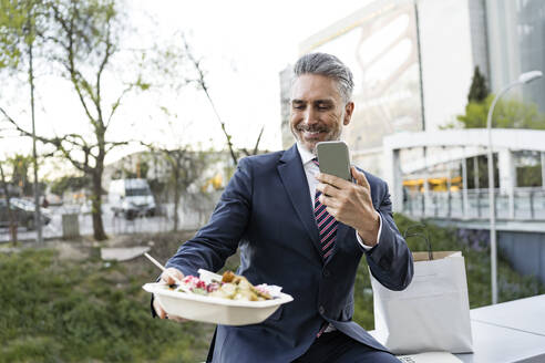 Geschäftsmann, der mit seinem Smartphone an der Wand sitzend eine Essensbox fotografiert - JJF00850