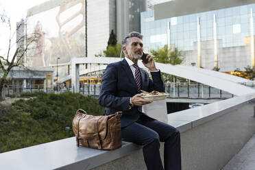 Businessman talking through smart phone holding meal sitting on wall - JJF00851