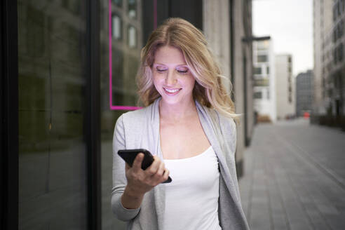 Smiling woman holding mobile phone at sidewalk - PNEF02689