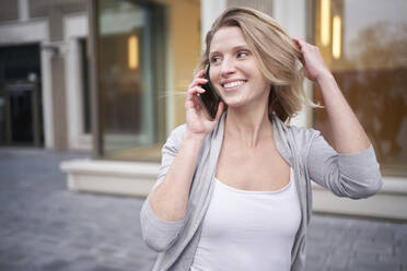 Junge lächelnde Frau mit Hand im Haar, die mit einem Smartphone spricht - PNEF02697