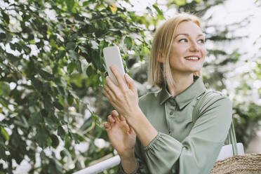 Happy woman with smart phone standing in greenhouse - NDEF00588