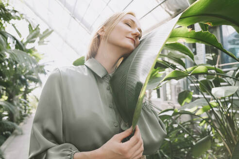 Smiling woman embracing banana leaf in green house - NDEF00597