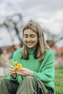 Lächelndes Teenager-Mädchen, das einen Kranz mit gelben Pusteblumen macht, die im Gras sitzen - VSNF00733