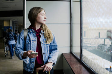 Junge Frau mit Smartphone schaut am Flughafen aus dem Fenster - JJF00857