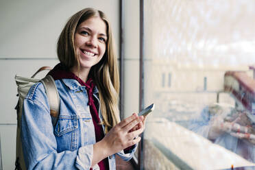 Smiling young woman holding smart phone by glass window at airport - JJF00859