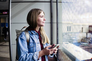 Junge Frau schaut aus dem Fenster und hält ihr Smartphone am Flughafen - JJF00860