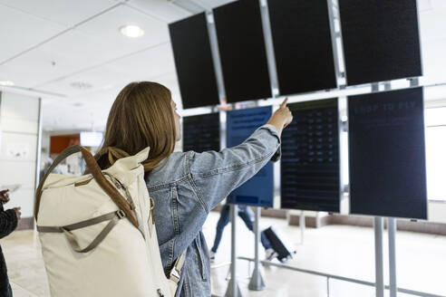 Woman with backpack pointing at arrival departure board - JJF00863