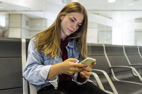 Junge Frau, die in der Lobby eines Flughafens eine SMS über ihr Smartphone verschickt - JJF00867