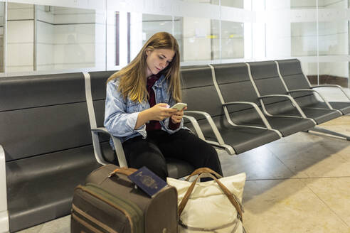 Junge Frau mit Smartphone auf dem Sitz in der Flughafenlobby - JJF00868