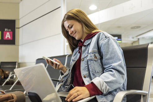 Lächelnde Frau mit Laptop und Smartphone in der Flughafenlobby - JJF00869