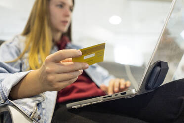 Junge Frau beim Online-Shopping mit Kreditkarte und Laptop in der Flughafen-Lobby - JJF00873