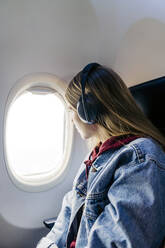 Frau mit Kopfhörern, die im Flugzeug sitzend aus dem Fenster schaut - JJF00880