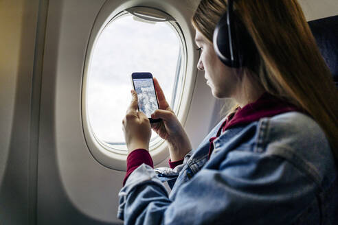 Frau mit Kopfhörern, die durch ein Flugzeugfenster mit ihrem Mobiltelefon fotografiert - JJF00881