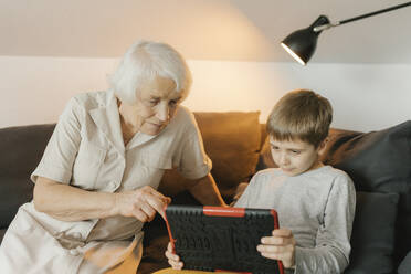 Boy sharing tablet PC with grandmother sitting on sofa at home - VIVF00591