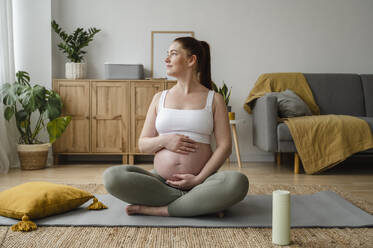 Schwangere Frau sitzt im Schneidersitz mit den Händen auf dem Bauch zu Hause - ALKF00197