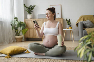 Lächelnde schwangere Frau, die ein Smartphone benutzt und zu Hause auf einer Übungsmatte sitzt - ALKF00201