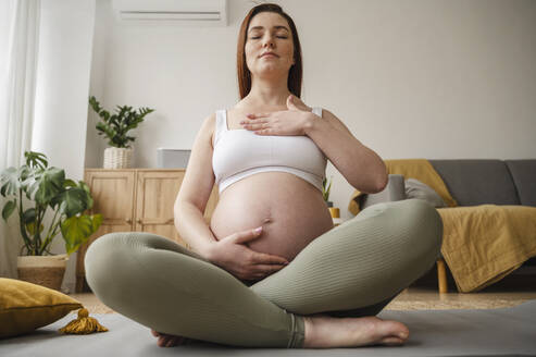 Schwangere Frau mit geschlossenen Augen sitzt im Schneidersitz auf einer Übungsmatte zu Hause - ALKF00203