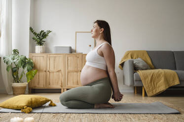 Schwangere Frau streckt die Hände auf einer Matte sitzend zu Hause - ALKF00208