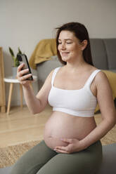 Lächelnde schwangere Frau, die ein Smartphone benutzt und zu Hause sitzt - ALKF00209