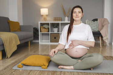 Schwangere Frau, die ihren Bauch berührt, sitzt im Schneidersitz auf einer Matte zu Hause - ALKF00212