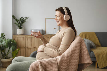 Lächelnde schwangere Frau, die Musik hört und ein Glas Wasser hält, während sie zu Hause sitzt - ALKF00219