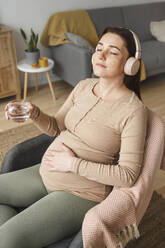 Schwangere Frau genießt Musik und hält ein Glas Wasser, während sie zu Hause sitzt - ALKF00220