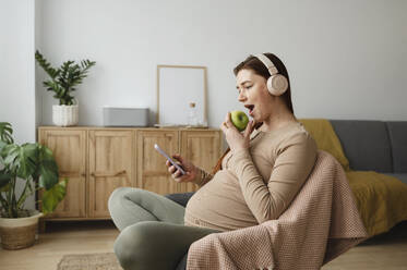 Schwangere Frau, die einen Apfel isst und ein Smartphone benutzt, während sie zu Hause auf einem Stuhl sitzt - ALKF00221