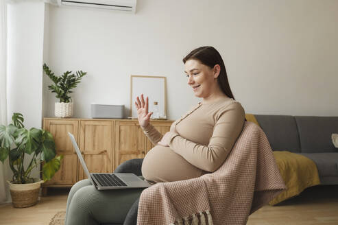 Lächelnde schwangere Frau winkt bei einem Videoanruf über einen Laptop zu Hause - ALKF00224