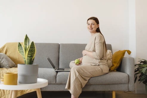 Lächelnde schwangere Freiberuflerin sitzt mit Laptop auf dem Sofa im Home Office - ALKF00228