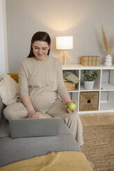 Smiling pregnant freelancer using laptop sitting on sofa at home office - ALKF00230