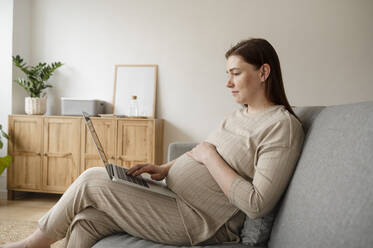 Pregnant freelancer with hand on stomach working on laptop at home office - ALKF00232