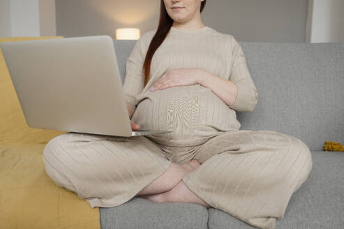 Schwangere Freiberuflerin mit Hand auf dem Bauch bei der Arbeit am Laptop im Home Office - ALKF00234