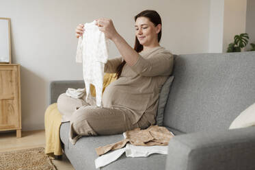 Lächelnde schwangere Frau mit Babykleidung, die zu Hause sitzt - ALKF00236