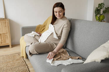 Lächelnde schwangere Frau mit Babykleidung sitzt zu Hause auf dem Sofa - ALKF00237
