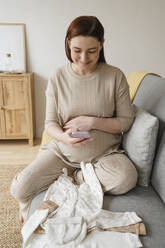 Lächelnde schwangere Frau, die ein Bild von Babykleidung auf dem Sofa zu Hause sitzt - ALKF00244