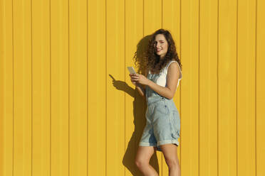 Smiling woman holding smart phone leaning on yellow wall - SYEF00367