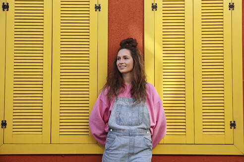 Lächelnde Frau in Latzhose steht an einer Wand mit gelben Fenstern - SYEF00368