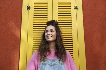 Lächelnde Frau mit langen Haaren in der Nähe einer Wand mit gelben Fenstern - SYEF00369