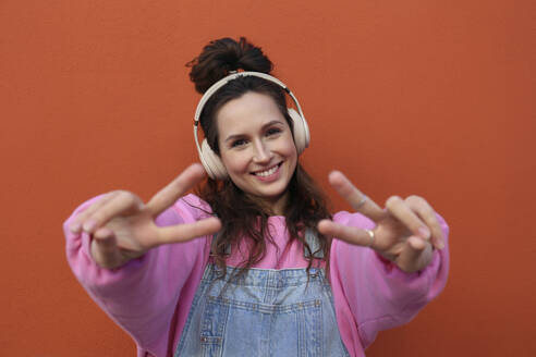 Happy woman showing peace sign wearing headphones against orange background - SYEF00372